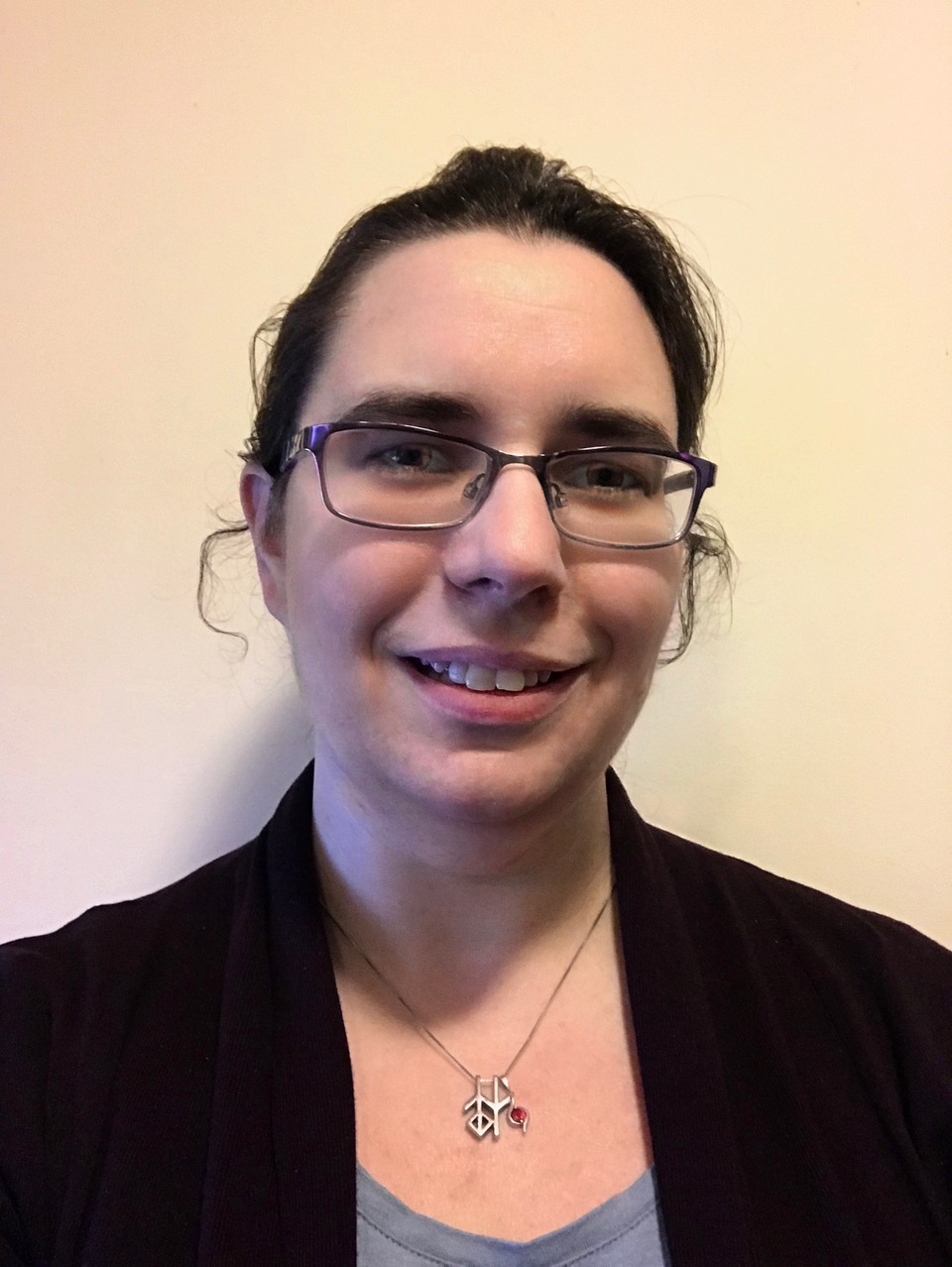 Smiling person with dark hair, purple glasses, and a silver pendant necklace on a light background.