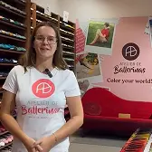 Woman wearing glasses and a white Atelier de Ballerinas t-shirt standing in a colorful shoe store with shelves of ballet flats and a red couch in the background.