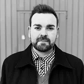 Portrait of a man with short hair, beard, and a cross earring wearing a checkered shirt, sweater, and dark jacket.