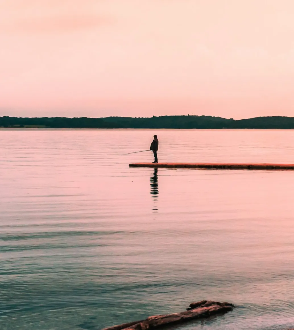 A person fishing while standing on a wooden pier in calm pink evening light.