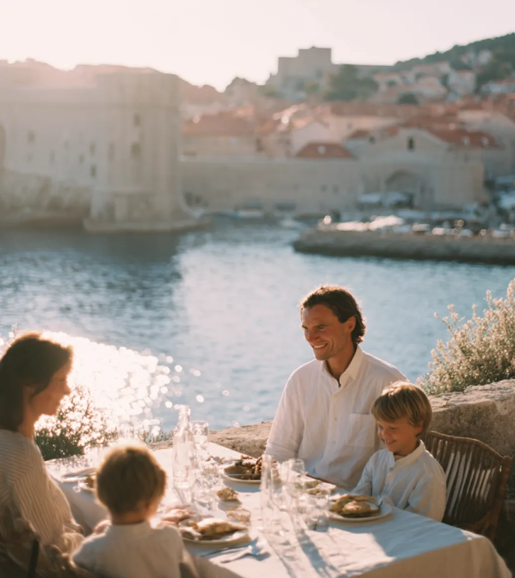 A family eating outdoors at a table near the water, smiling in the sunshine.