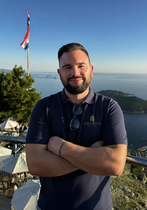 A smiling man in a white shirt standing against a light wooden wall.