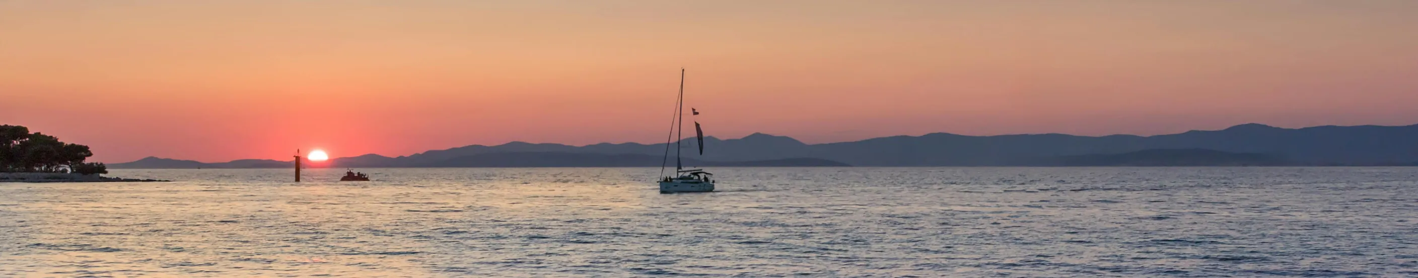 Sunset over mountains with a sailboat silhouette on calm water.