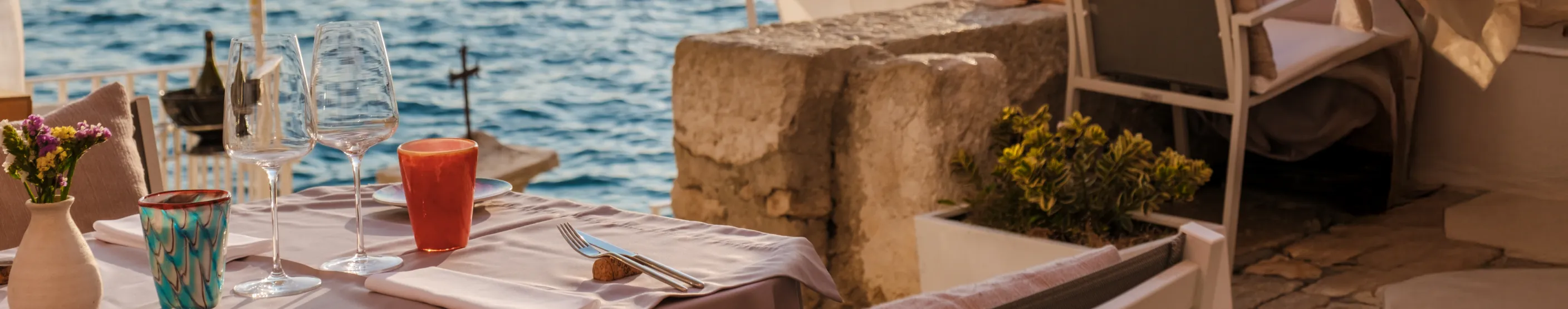 Beachfront dining table set with glasses and colorful cups beside the sea.
