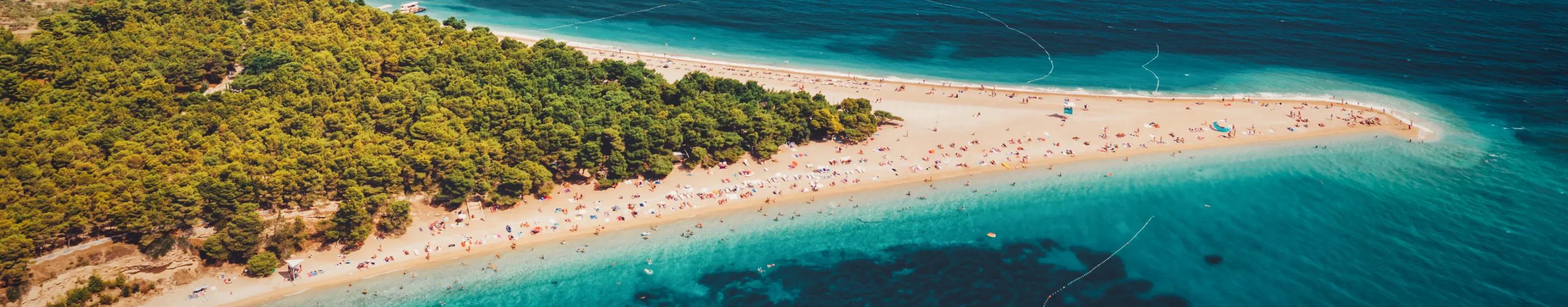 Aerial view of a narrow sandy beach surrounded by turquoise water and pine trees.