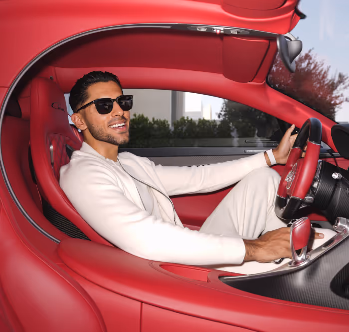 Man wearing sunglasses and white outfit sitting inside a luxury red sports car, smiling and holding the steering wheel.