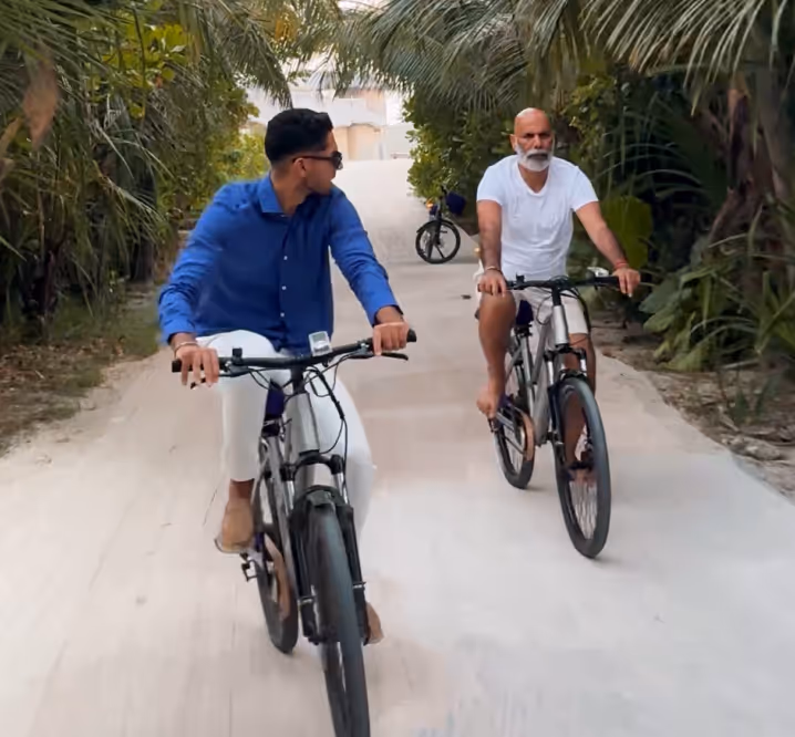 Two men riding bicycles barefoot on a sandy path lined with tropical palm trees.