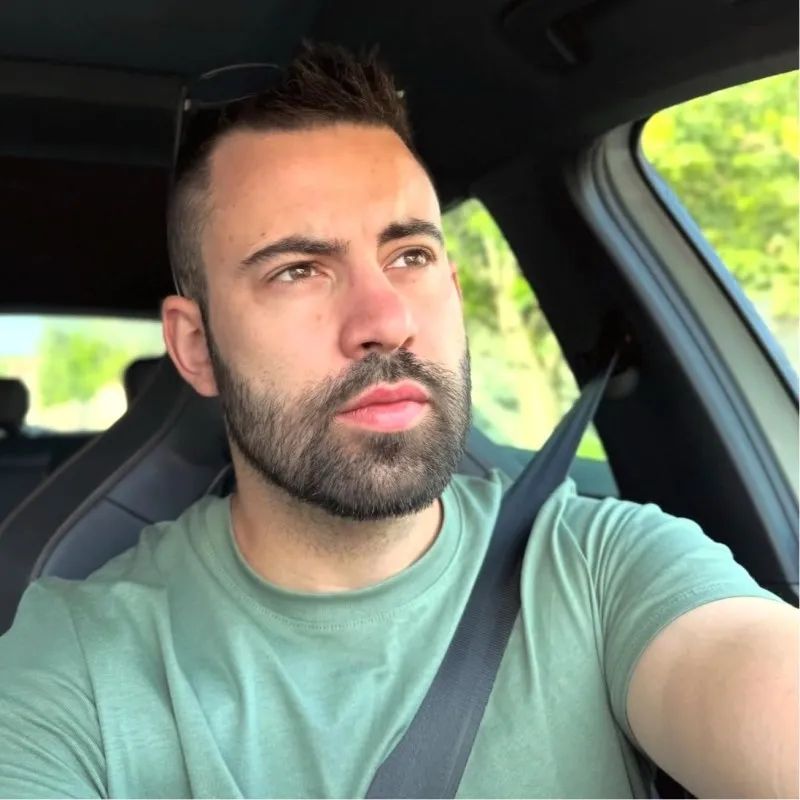 Homme barbu portant un t-shirt vert et une ceinture de sécurité, assis dans une voiture avec de la verdure visible à travers la fenêtre.