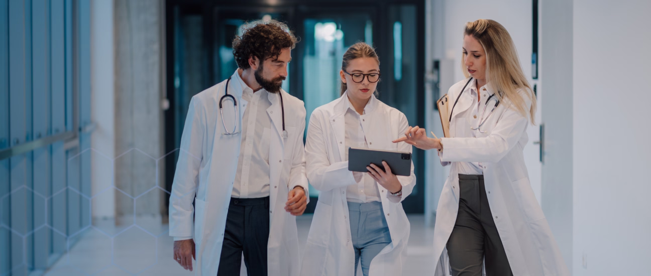 Three doctors in white coats walking down a hospital corridor discussing information on a digital tablet.