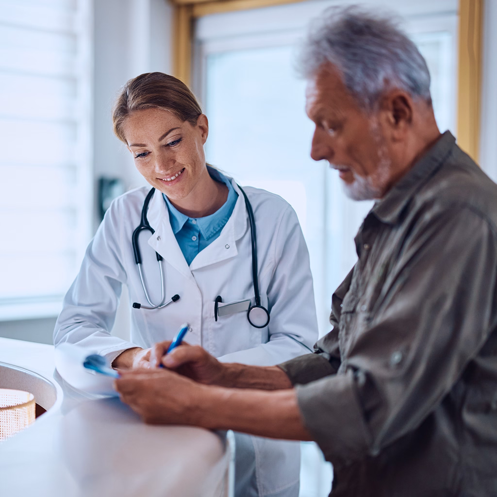 Female doctor with stethoscope smiling while consulting an elderly male patient who is writing.