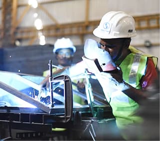 Worker wearing a white hard hat and reflective vest welding metal in an industrial setting.