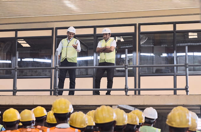Two safety inspectors wearing white helmets and yellow vests addressing a group of construction workers wearing yellow helmets.