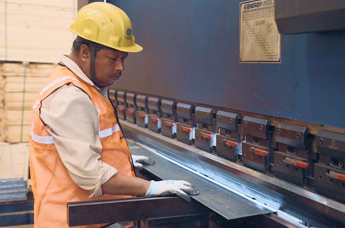 Factory worker in a yellow hard hat and orange safety vest operating a metal bending machine.