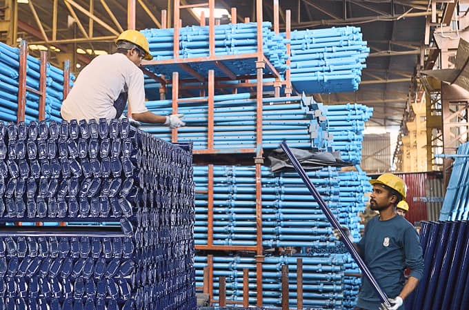 Two workers wearing yellow helmets handling blue metal pipes in an industrial warehouse with stacked pipes on racks.