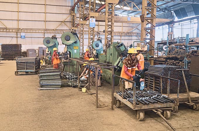 Workers in yellow helmets and orange uniforms operating machinery and handling metal components inside a spacious industrial factory.