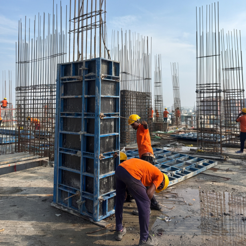 Construction workers in orange shirts and yellow helmets working with steel reinforcements and formwork on a building site under a clear sky.