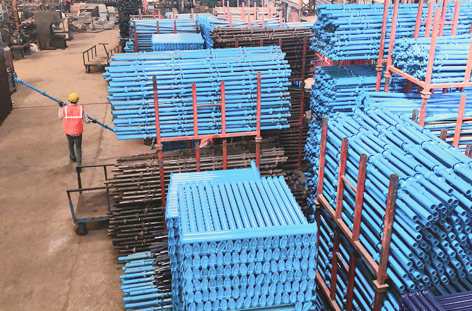 Warehouse with stacks of blue and brown metal pipes and a worker in a yellow helmet and orange vest pulling a cart.