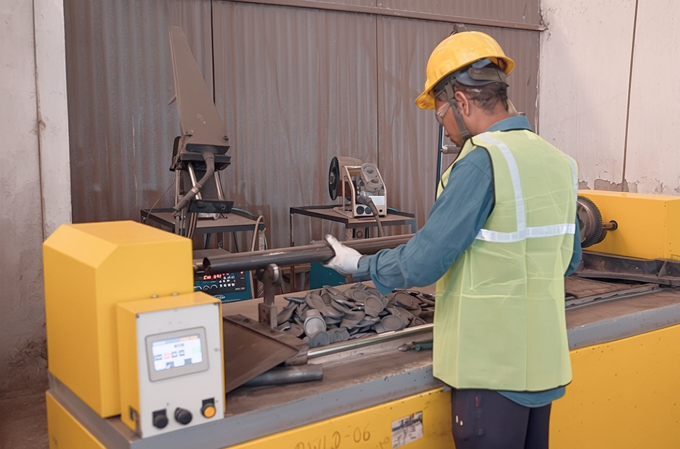 Worker in yellow hard hat and safety vest operating a yellow industrial machine handling metal pipes in a factory setting.