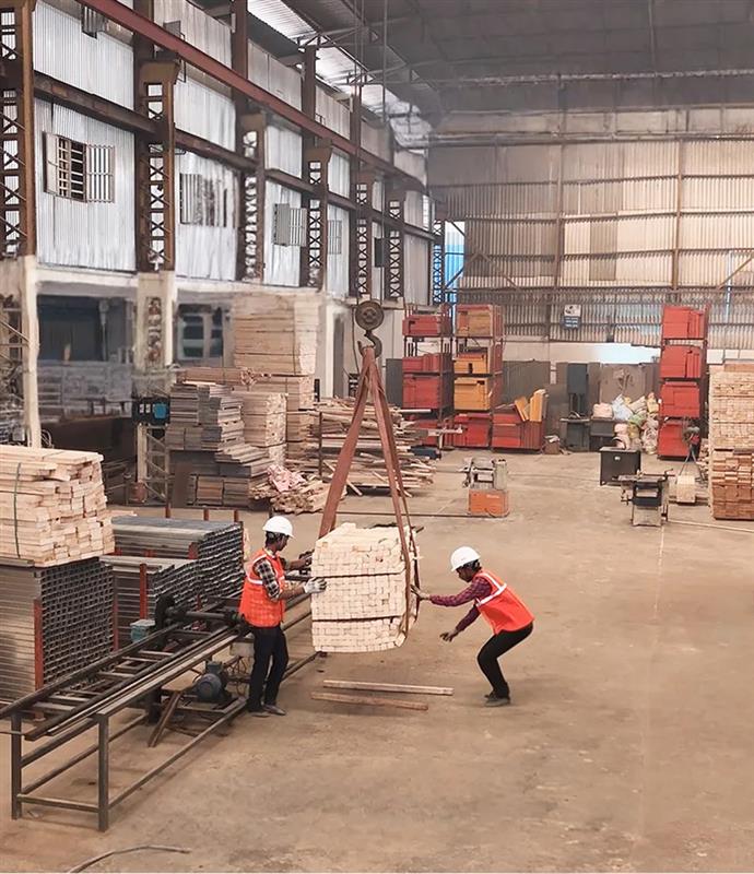 Two workers wearing white helmets and orange safety vests handling a large bundle of wooden planks suspended by a crane inside a spacious warehouse.