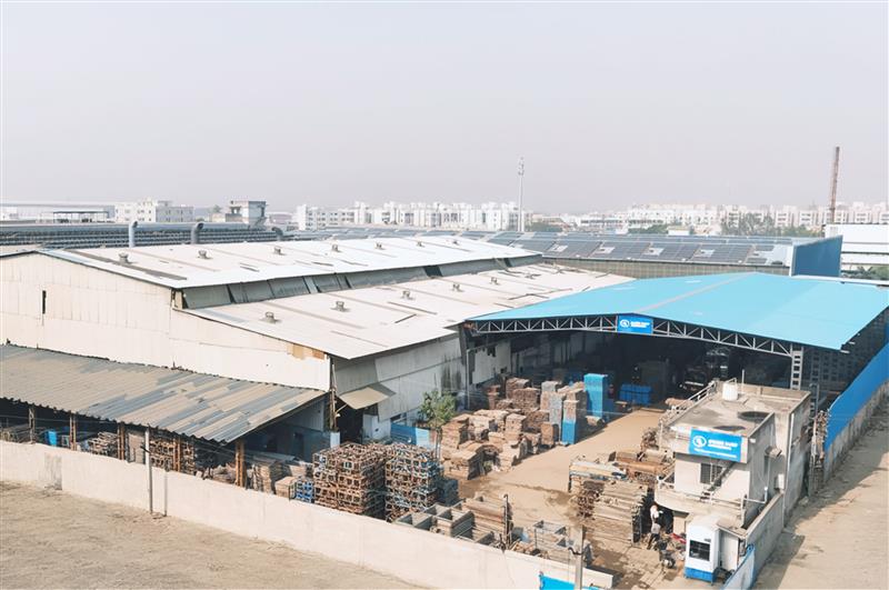 Aerial view of an industrial warehouse complex with multiple metal-roofed sections, stacks of pallets and crates in an open yard, and a small building with a blue sign at the entrance.