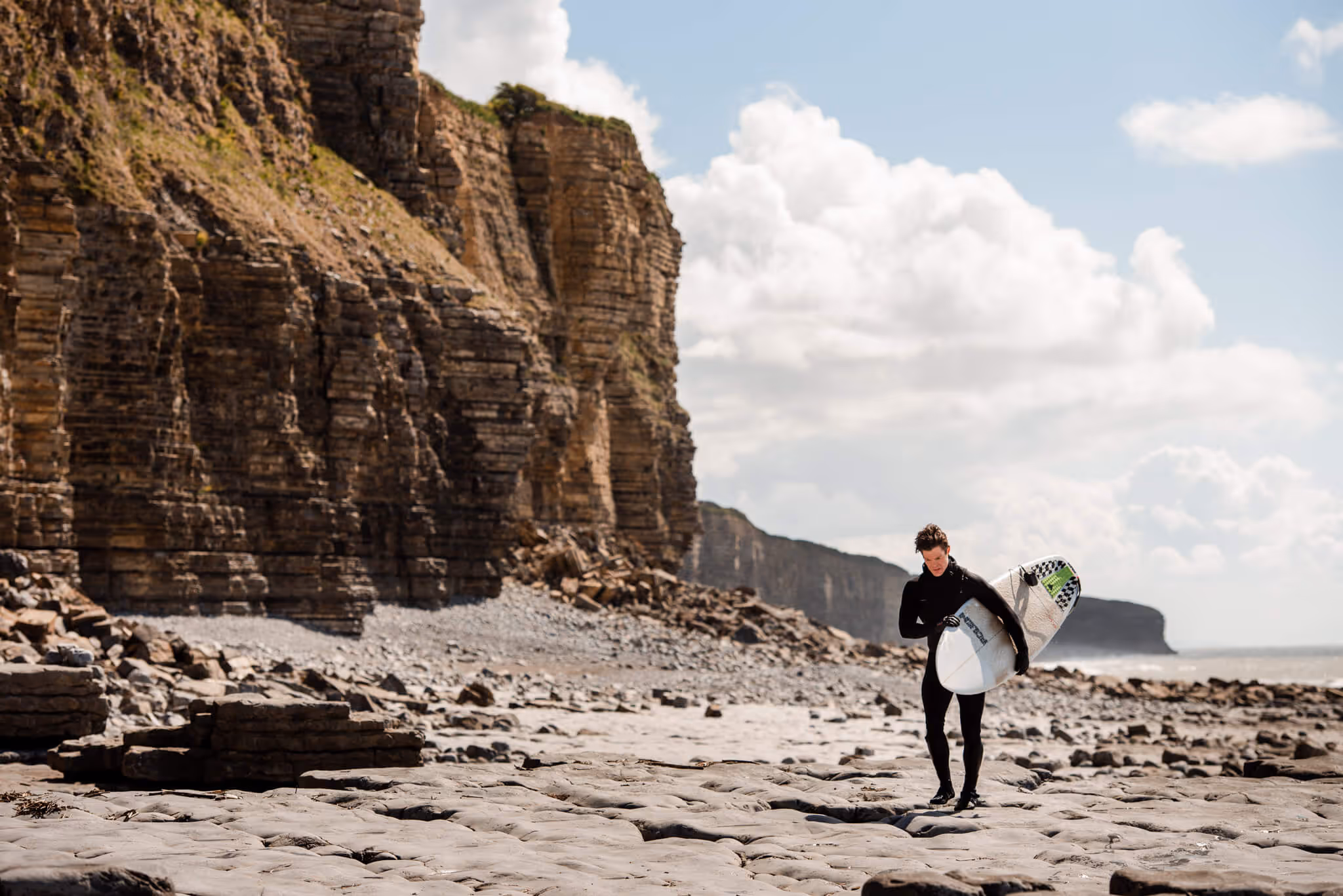 Surfer in a black wetsuit carrying a surfboard walking on rocky beach with large cliffs in the background under a partly cloudy sky.