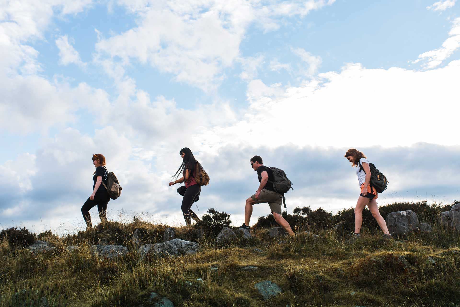 Four hikers walking single file along a grassy hill under a cloudy blue sky.