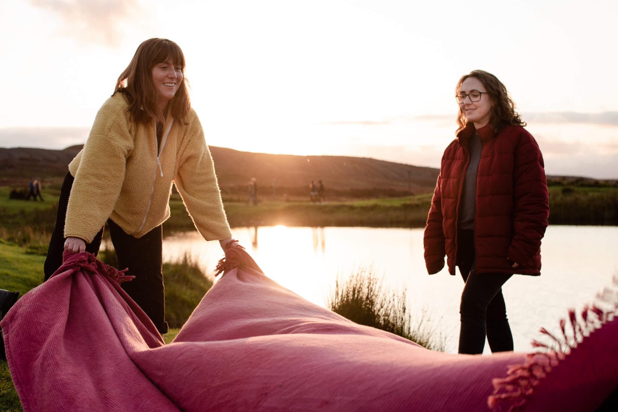 Two women outdoors near a pond at sunset, unfolding a large pink blanket.