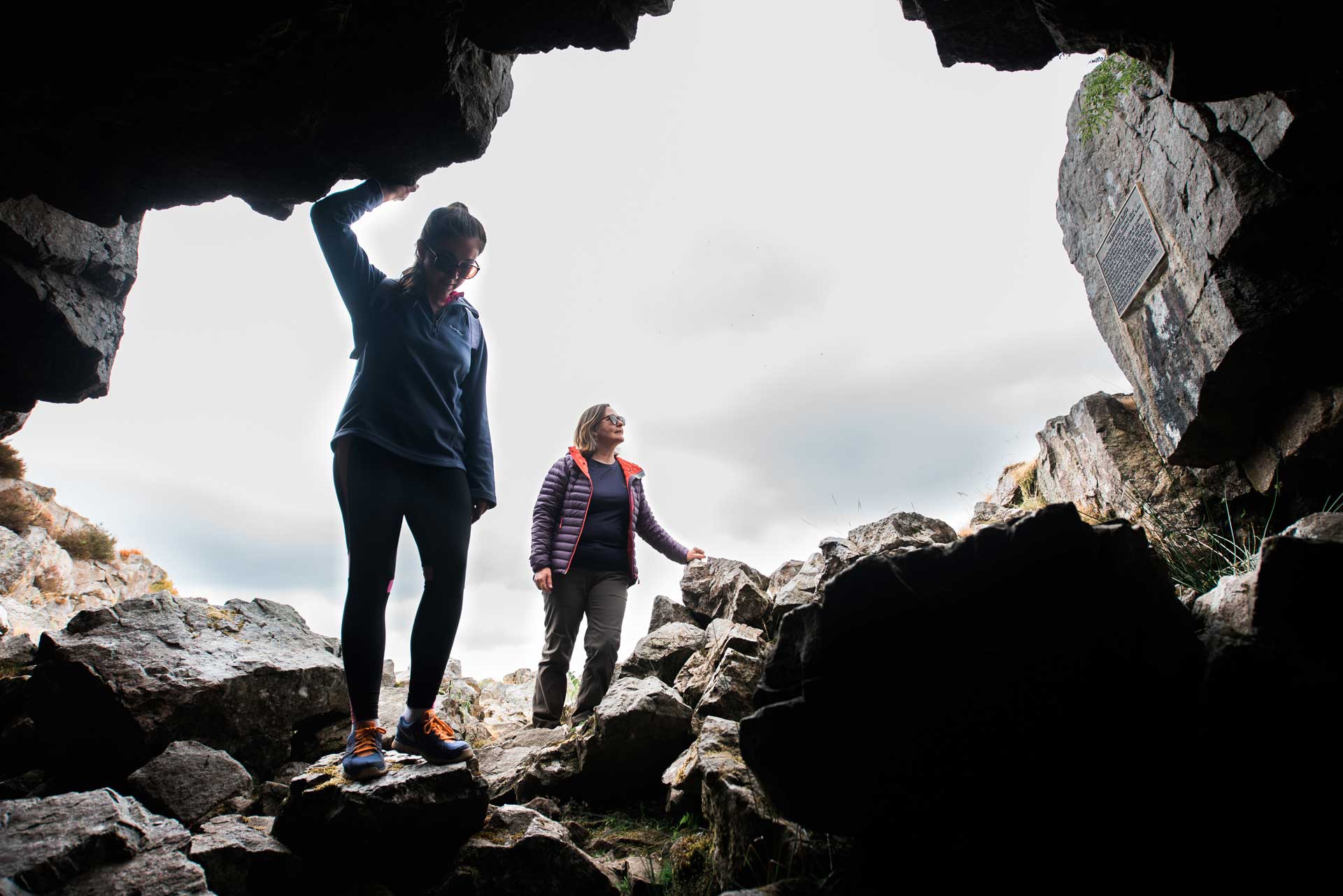 Two women hiking on rocky terrain, viewed from inside a cave opening.