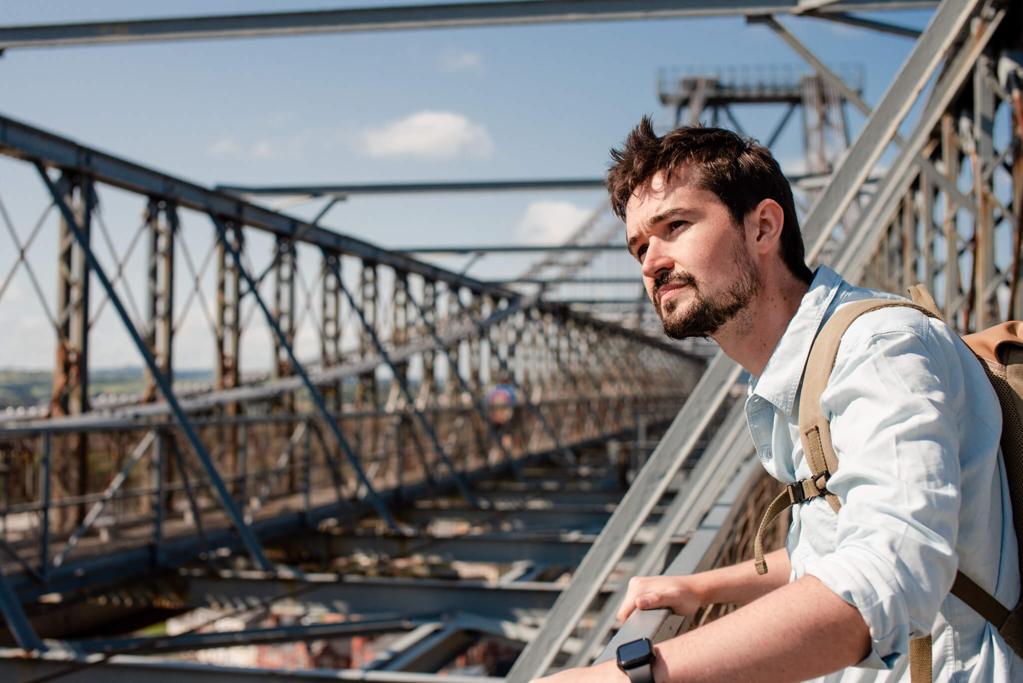 Man with a beard and backpack leaning on a railing on a large metal bridge under a blue sky.