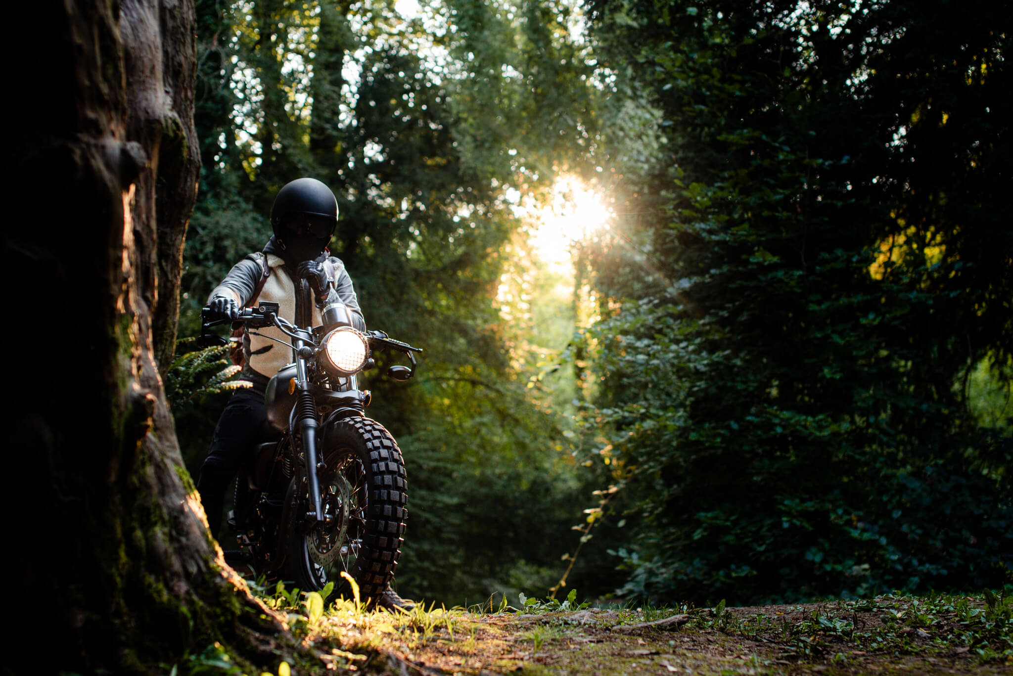 Motorcyclist wearing a black helmet and gloves riding a bike with a lit headlight on a forest path at sunset.