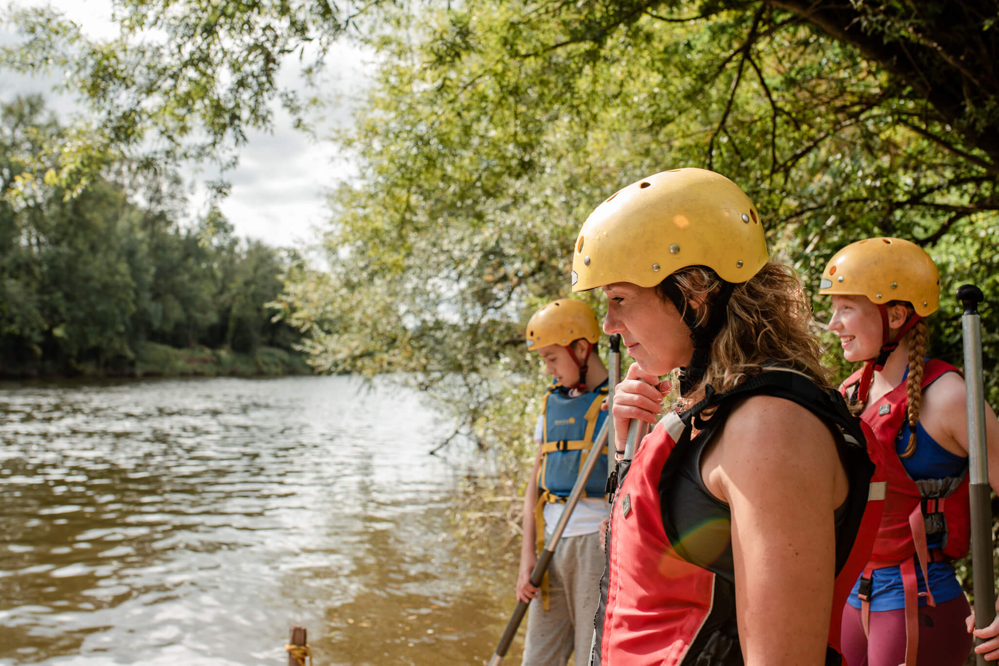 Three people wearing yellow helmets and red life jackets standing by a river holding paddles, surrounded by green trees.