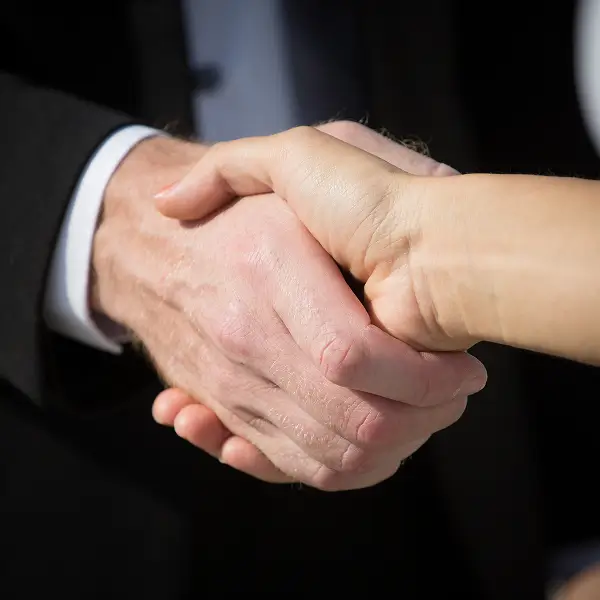 Close-up of two people shaking hands, one wearing a black suit with a white shirt.