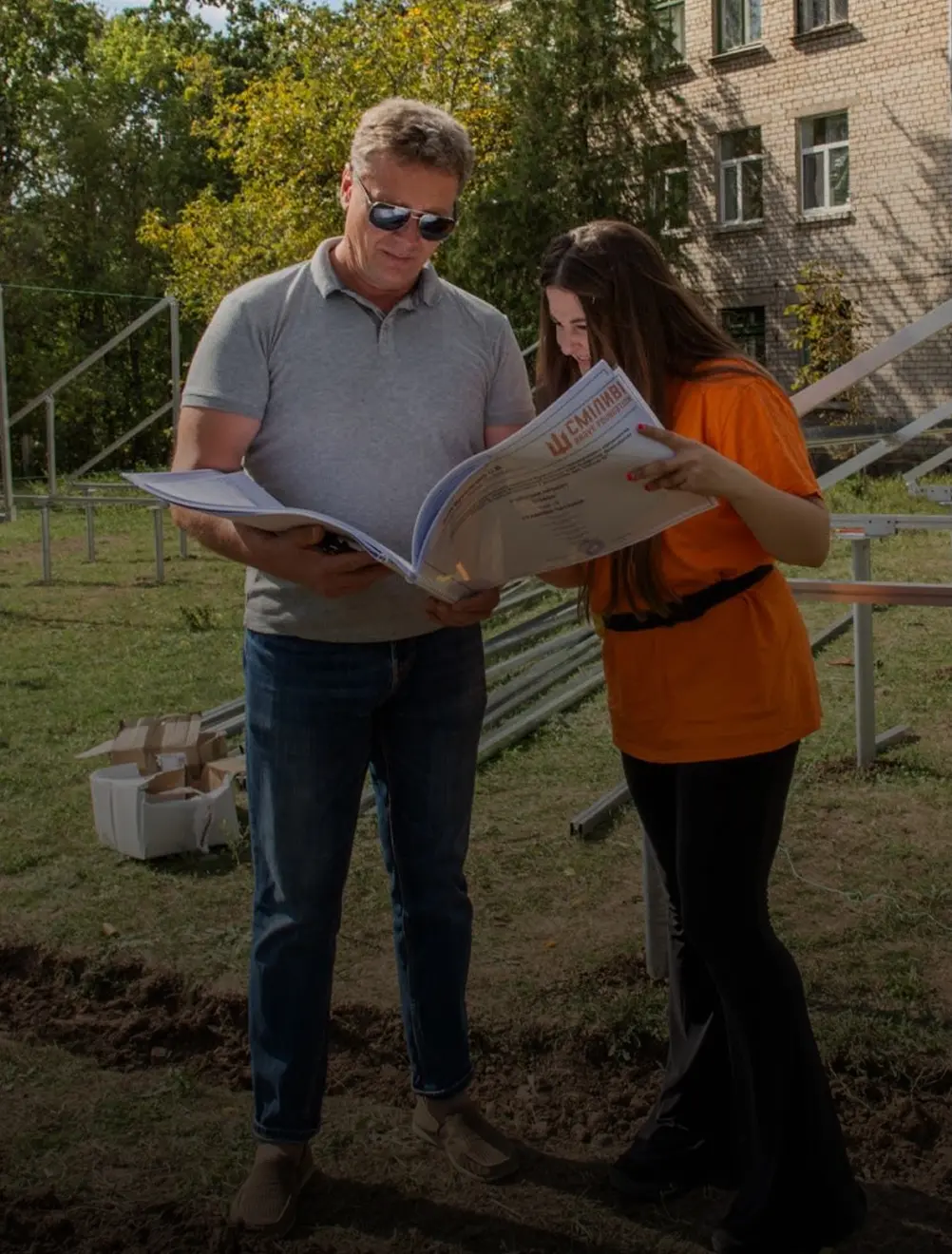A man in sunglasses and a gray polo shirt and a woman in an orange shirt studying a large document together outdoors.