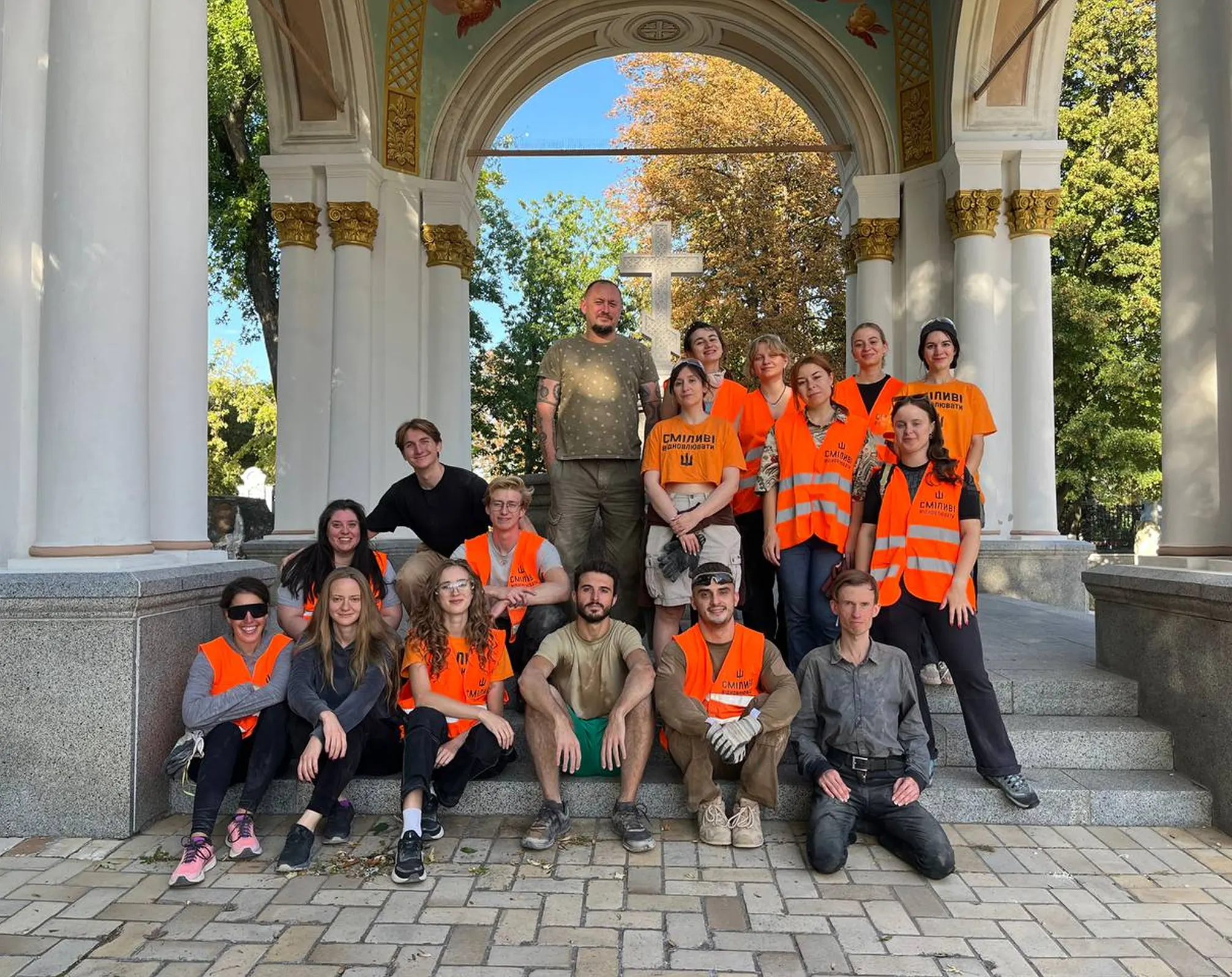 Group of people wearing orange safety vests and casual clothing posing on stone steps under a decorated arch with a cross and trees in the background.