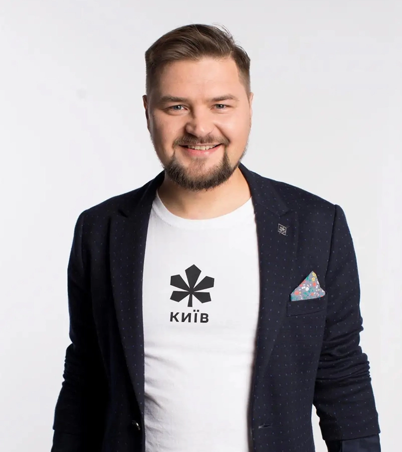 Smiling man with beard wearing a white t-shirt with a black symbol and 'Київ' text under a dark blazer with a patterned pocket square.