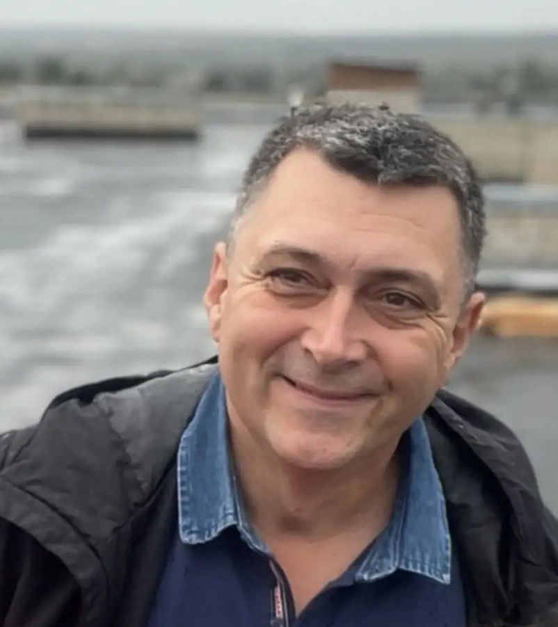 Smiling middle-aged man with short graying hair wearing a blue denim shirt and black jacket outdoors.