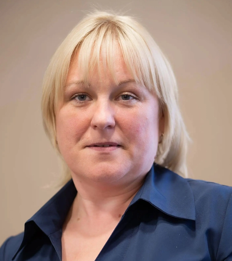 Portrait of a woman with blonde hair and blue eyes wearing a dark blue collared shirt against a plain beige background.