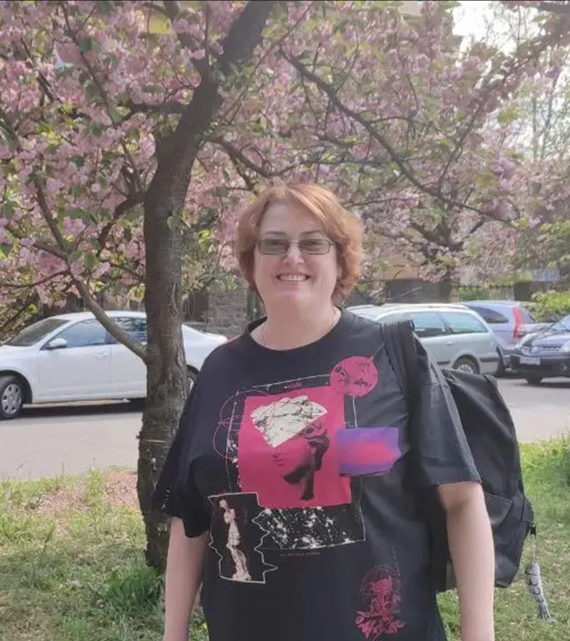 Smiling person with short light brown hair, glasses, and black graphic t-shirt stands outdoors with a black backpack and pink flowering tree in the background.