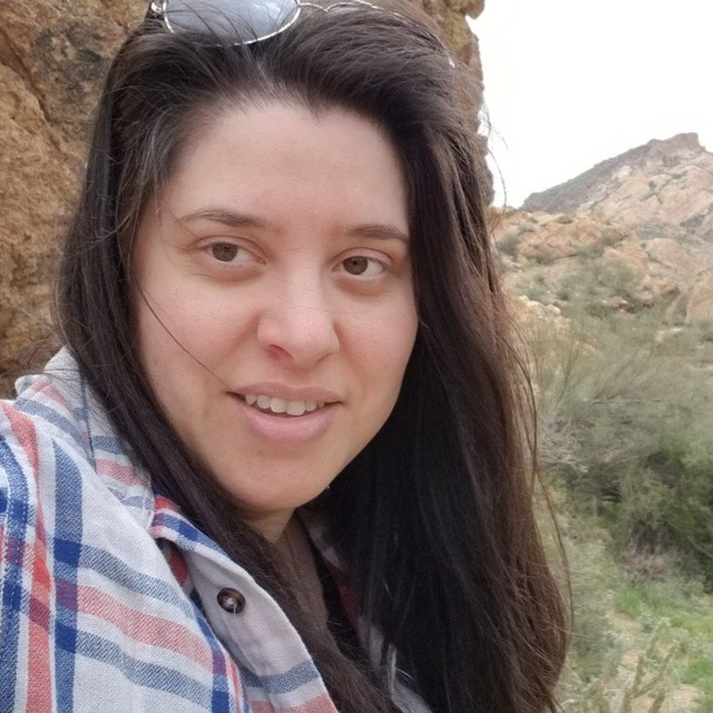 Woman with long dark hair wearing a plaid shirt outdoors near rocky terrain and greenery.