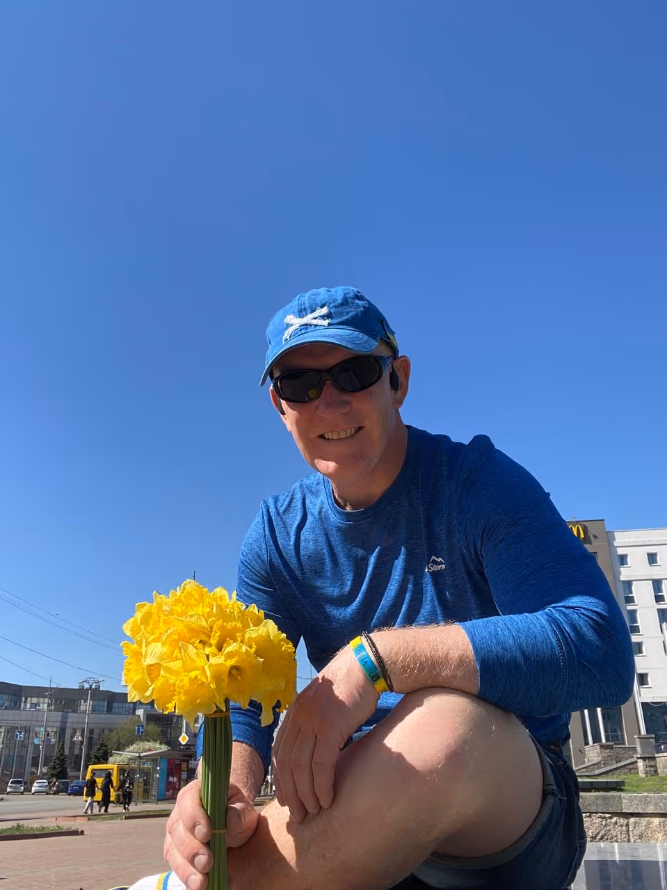 Man wearing sunglasses, a blue cap and blue long-sleeve shirt, holding a large bunch of yellow daffodils outdoors on a sunny day.