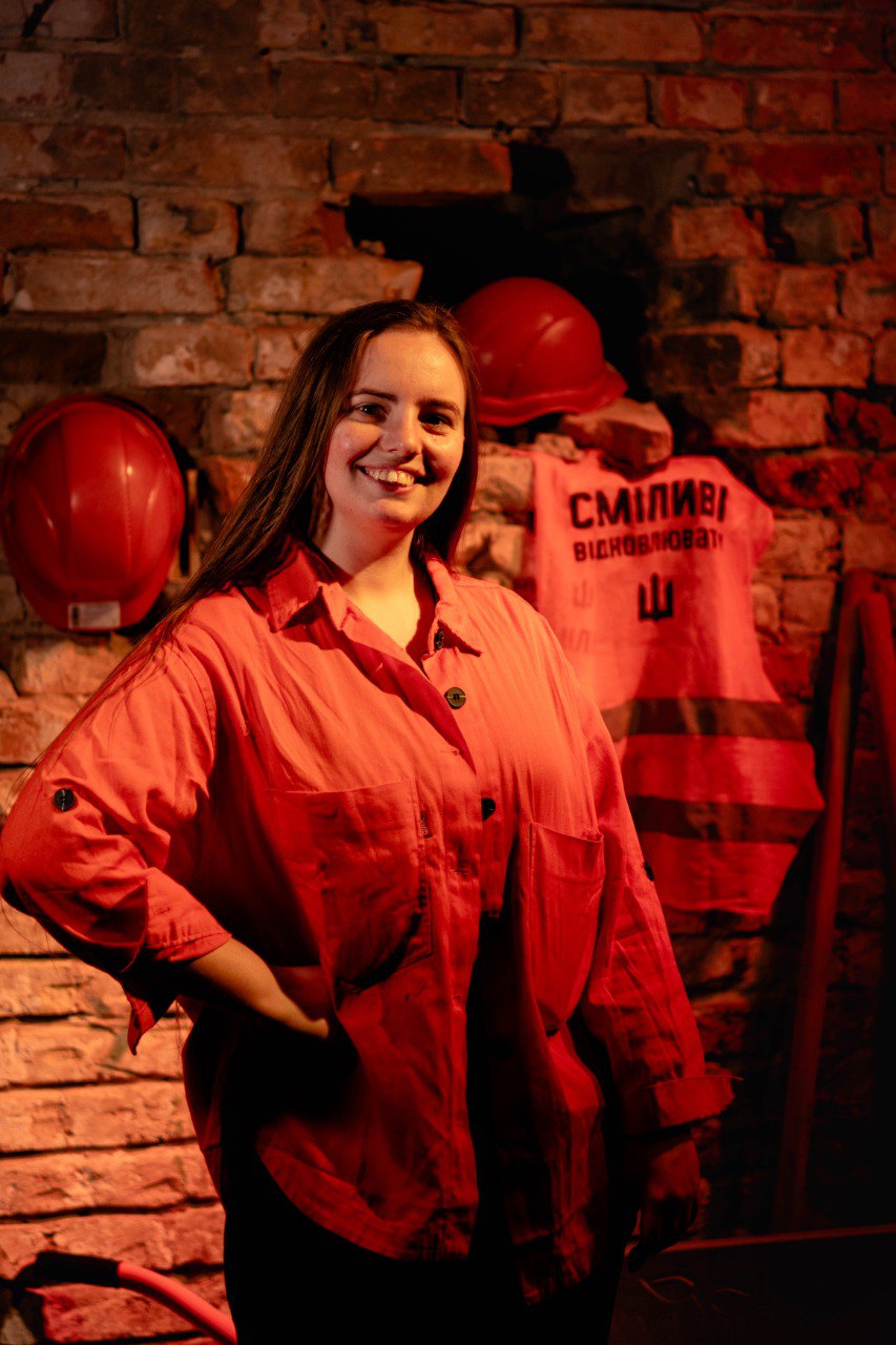 Smiling woman in a red jacket standing in front of a brick wall with red safety helmets and a reflective vest hanging behind her.