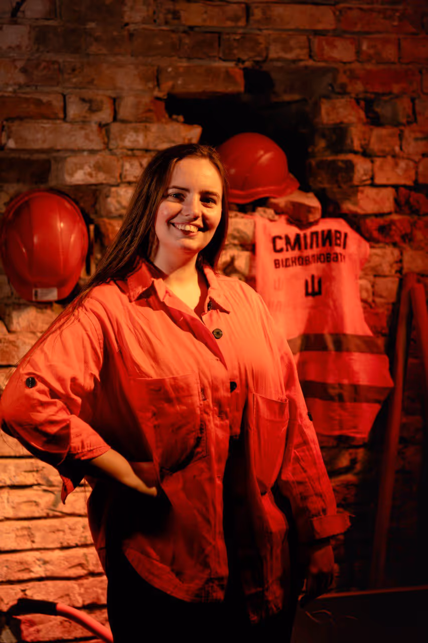 Smiling woman in a red jacket standing in front of a brick wall with red safety helmets and a reflective vest hanging behind her.