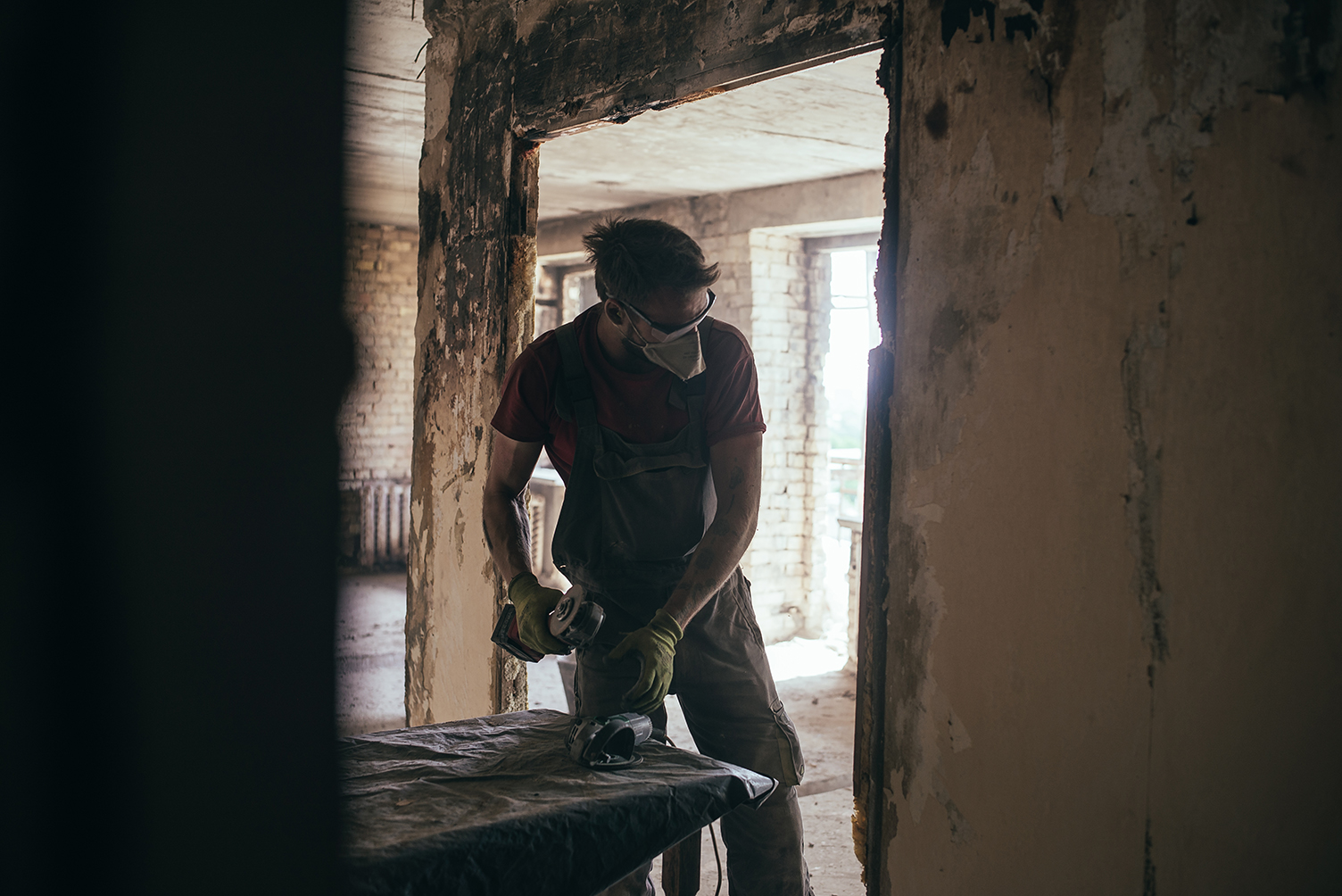 Construction worker wearing a protective mask and gloves using an angle grinder inside a partially built or renovated room.