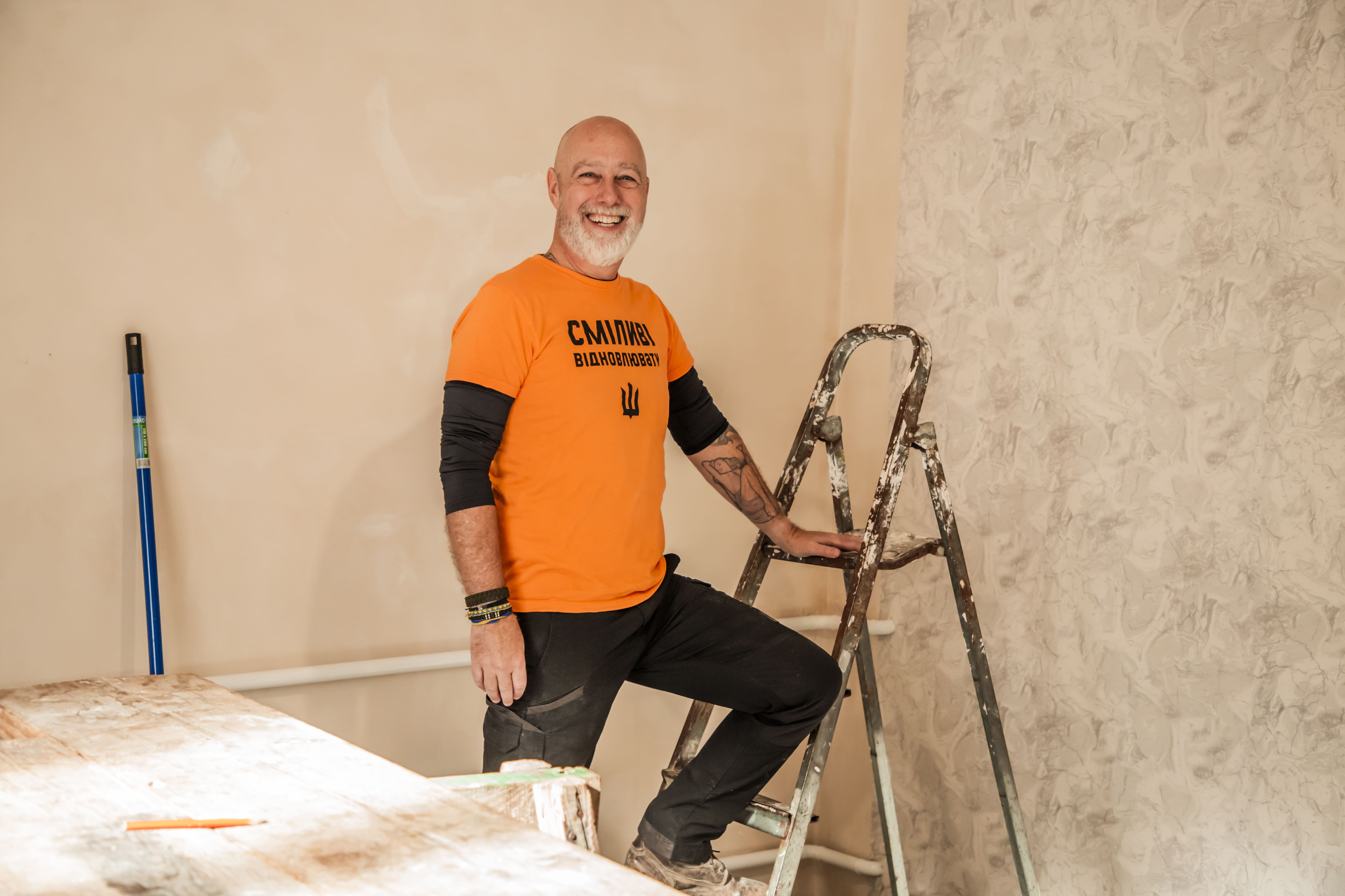Smiling man in an orange shirt and black pants standing on a paint-spattered ladder inside a room under renovation.