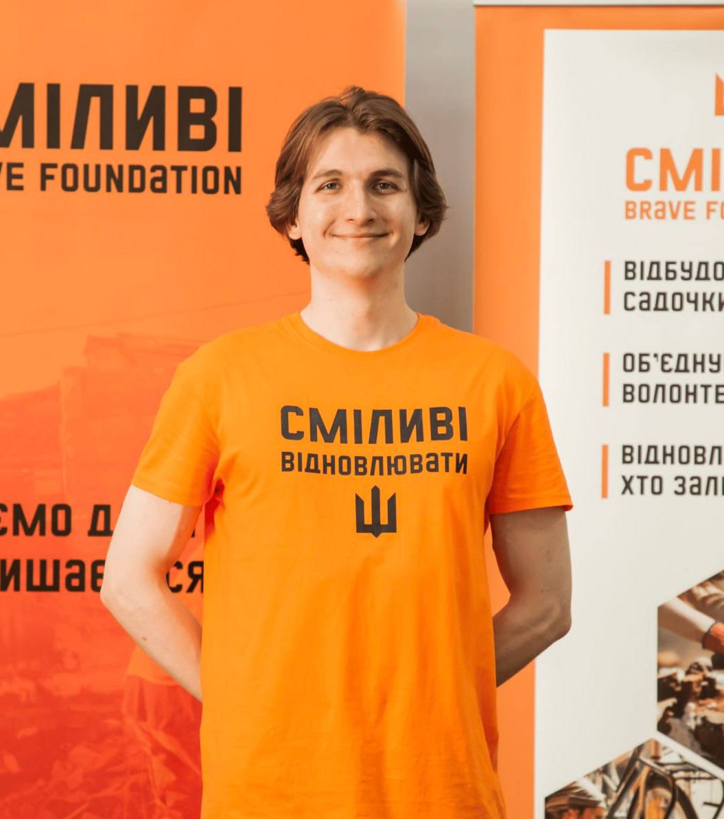 Young man smiling and wearing an orange t-shirt with Ukrainian text standing in front of orange and white banners with similar text.