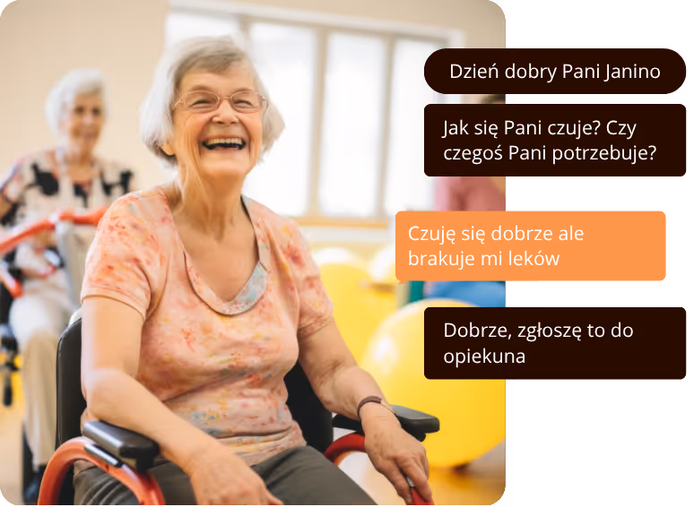 Starsza kobieta uśmiecha się siedząc na wózku inwalidzkim w jasnym pomieszczeniu, w tle inne osoby i kolorowe piłki.