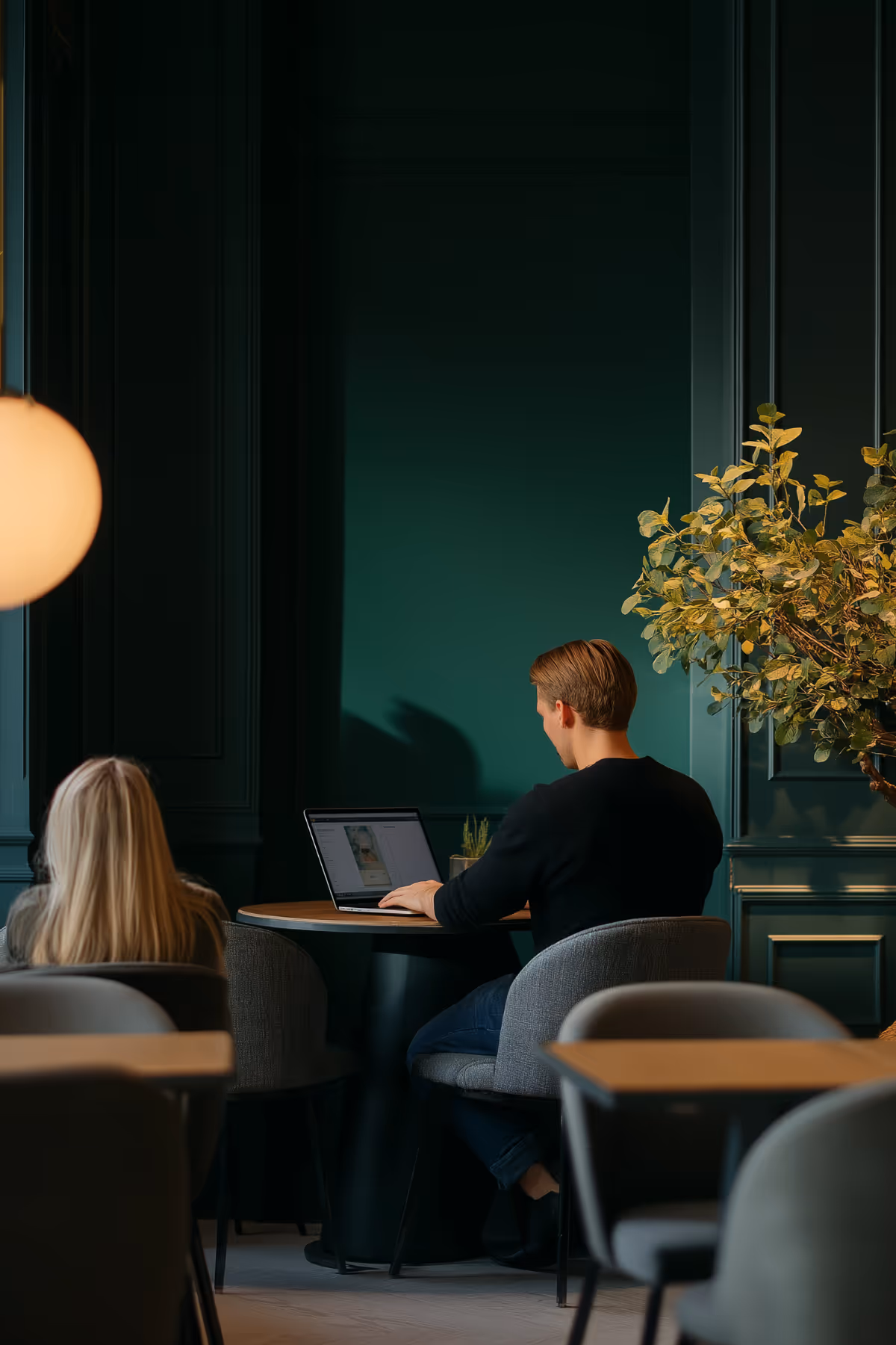 Man working on a laptop at a round table in a dimly lit room with teal walls and a large leafy plant nearby.