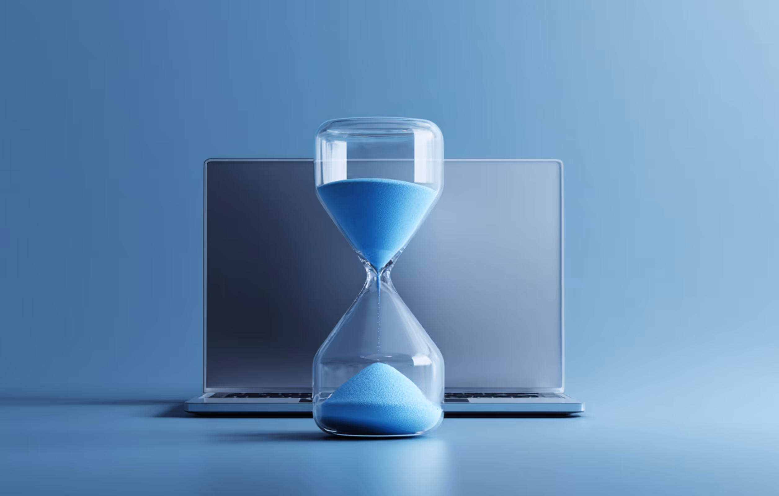 Transparent hourglass with blue sand in front of a closed laptop on a blue surface.