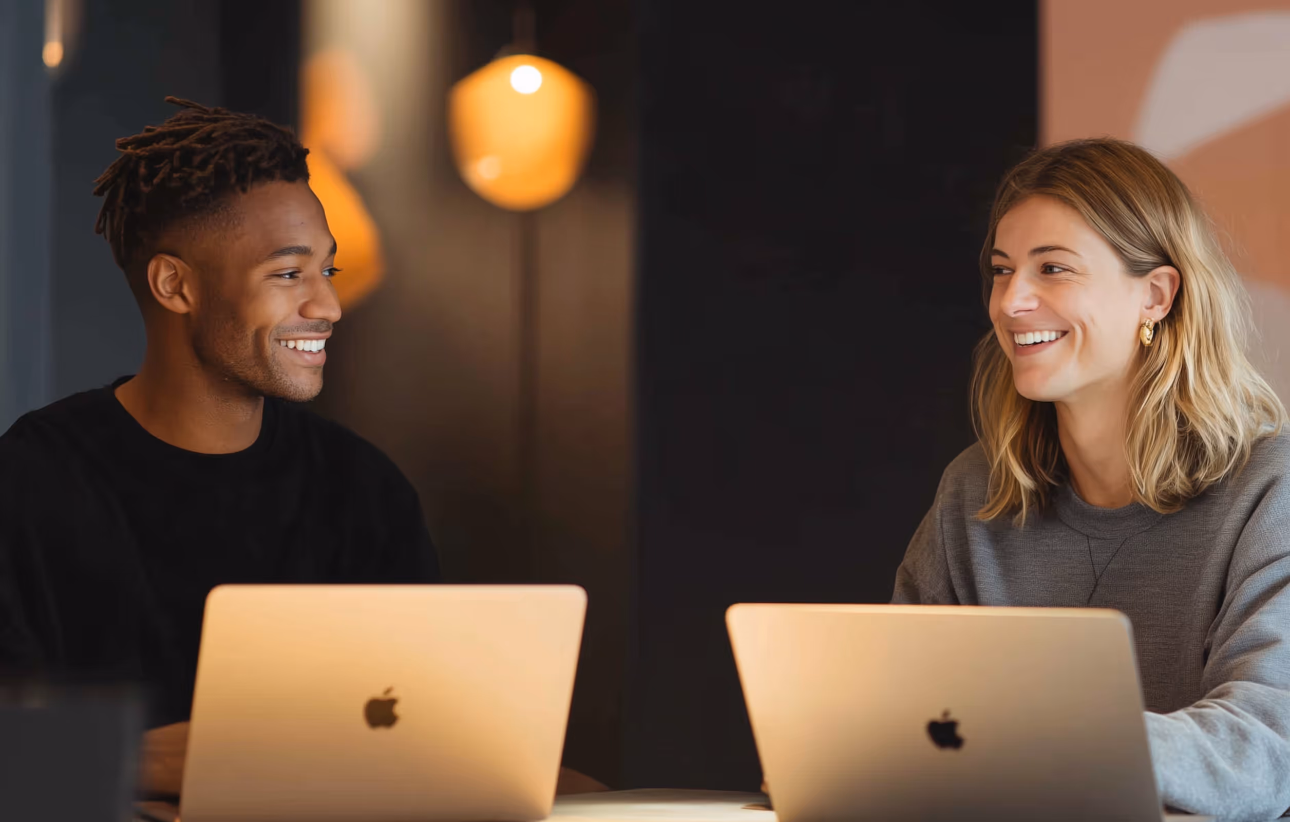 Two people smiling at each other while sitting at a table with Apple laptops in a warmly lit room.
