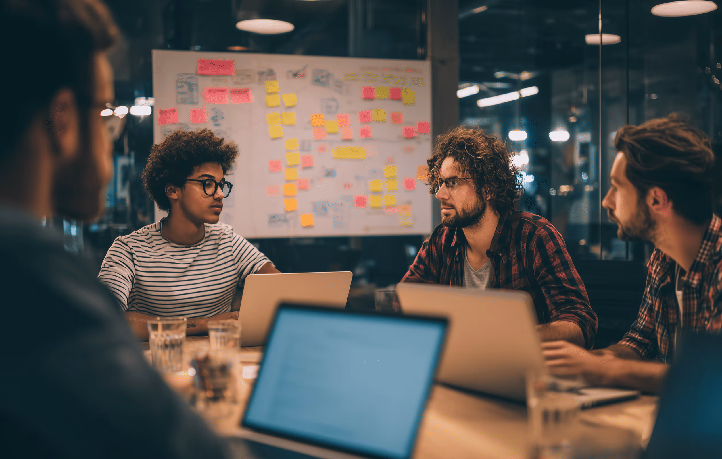 Four young professionals in a meeting room with laptops, discussing in front of a board covered with colorful sticky notes.